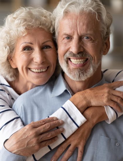 Head shot portrait of affectionate loving middle aged hoary beautiful woman cuddling from back smiling old husband. Happy loving mature married family couple looking at camera, posing for photo.